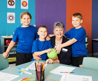 Children in blue shirts pose with a smiling elderly woman in a colorful classroom with superhero posters.