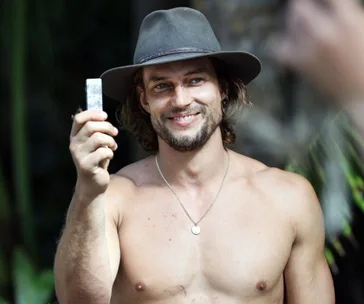 Shirtless man with a hat smiling and holding a small, shiny object outdoors.