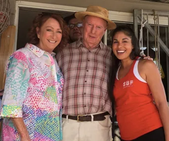 Three people smiling together, one in a colorful shirt, one in plaid with a hat, and another in a red tank top.