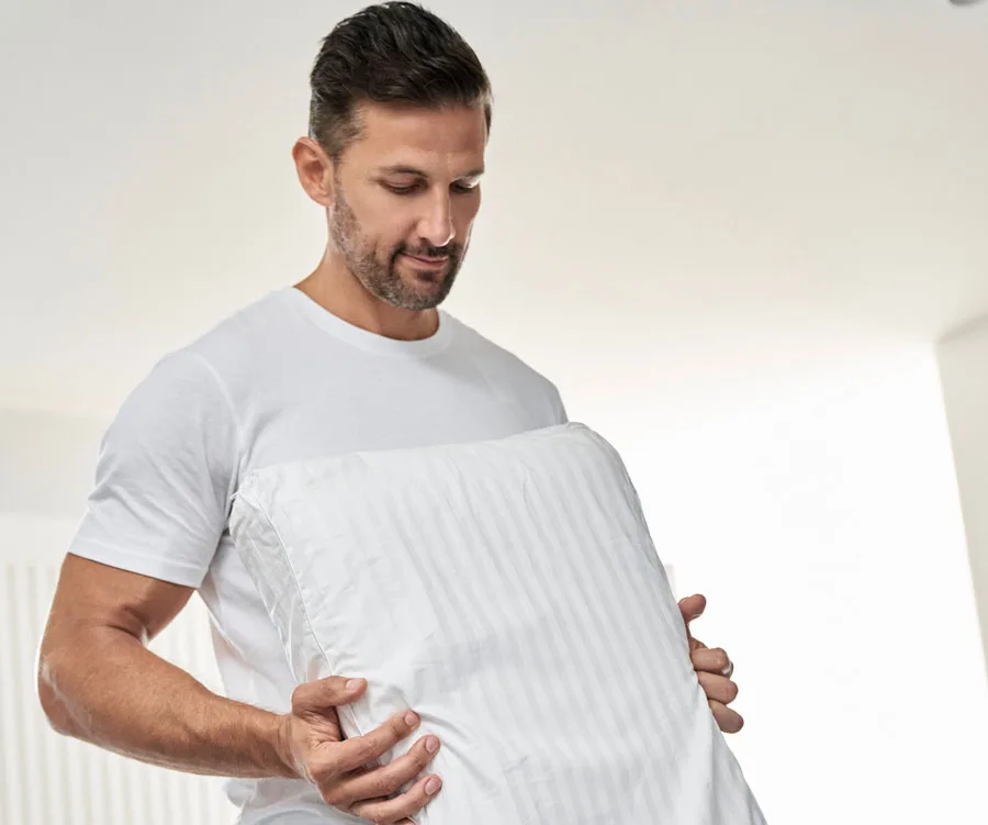 A person in a white shirt holding a white pillow in a bright room.