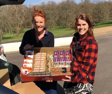 Two women smiling and holding a box of groceries while standing near an open car trunk in a sunny outdoor setting.