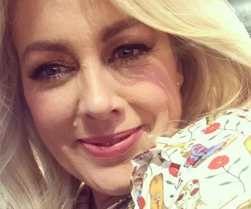Blonde woman smiling, wearing a floral patterned shirt, close-up view with soft lighting.