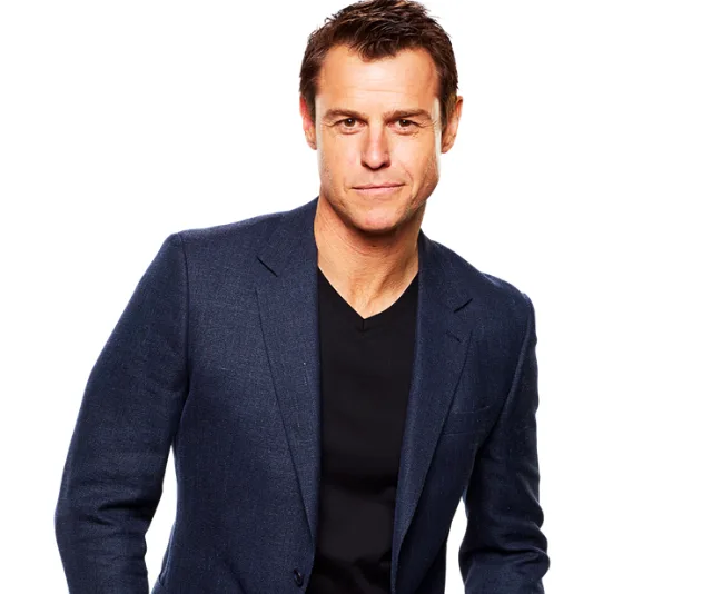Man in a navy blazer and black shirt against a white background, appearing confident.