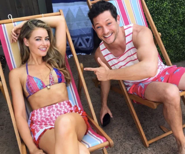 "Two people smiling on beach chairs, one pointing at the other, wearing colorful summer outfits."