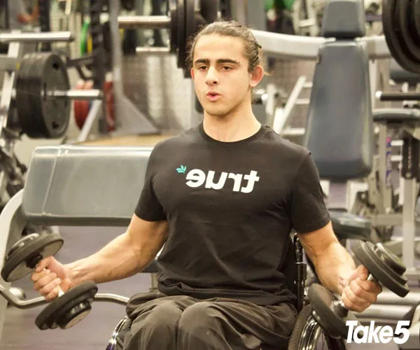 Man in a wheelchair lifting dumbbells at the gym, wearing a "true" t-shirt.