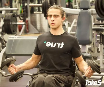Man in a wheelchair lifting dumbbells at the gym, wearing a "true" t-shirt.