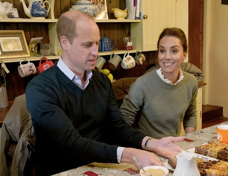 Two people sitting at a table with mugs, cakes, and decorative teapots on a shelf behind them.