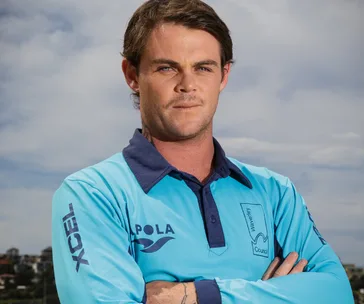 A lifeguard in a blue uniform poses with arms crossed, background shows a cloudy sky and distant shoreline.