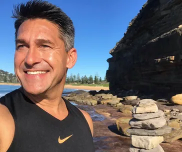 Man smiling outdoors near a rocky coastline with blue sky and stone cairn.