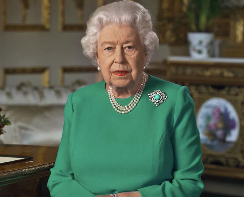 Queen Elizabeth II delivering a speech, wearing a green dress with pearl necklace and brooch, ornate room background.