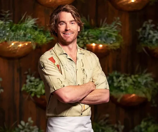 A man with curly hair smiles, wearing a patterned shirt and apron, standing in front of hanging plants.