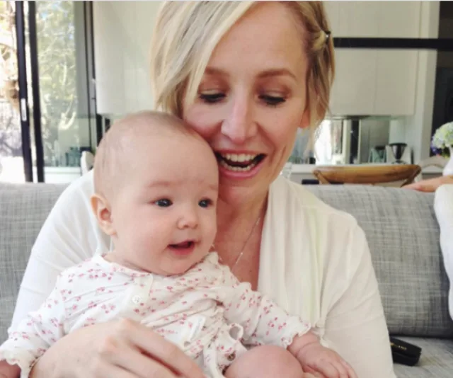 A woman smiles while holding a baby in a cozy living room setting.