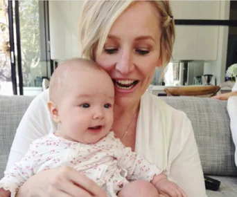 A woman smiles while holding a baby in a cozy living room setting.