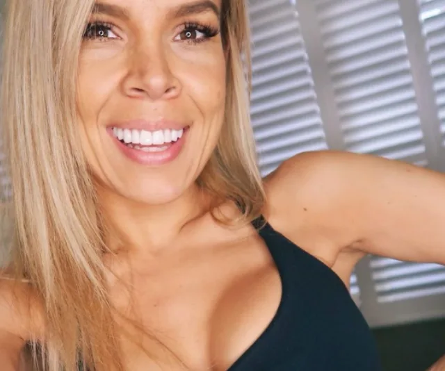 Smiling woman with long blonde hair and black tank top, standing in front of window blinds.