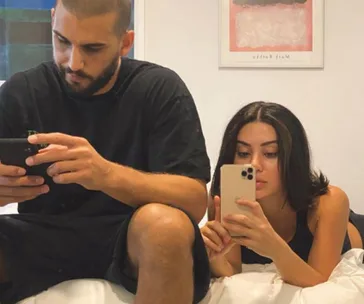 A couple sitting closely on the bed, focused on their smartphones, with a framed art piece hanging on the wall behind them.