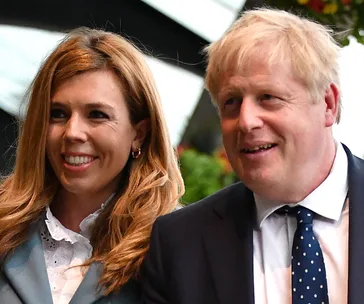 Two individuals, one with long hair and one with short hair, dressed in formal attire, standing outdoors and smiling.