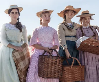 Four women in period dresses and straw hats hold baskets, standing outdoors with a sunny sky.