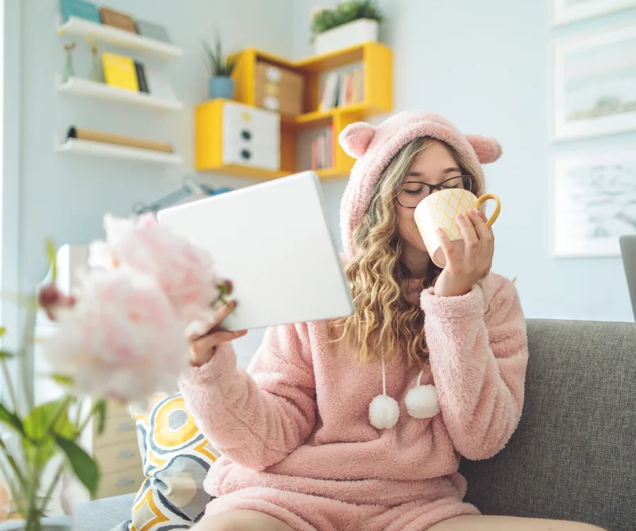 Woman in pink cozy outfit with bear ears, holding a tablet and sipping from a mug on a sofa.
