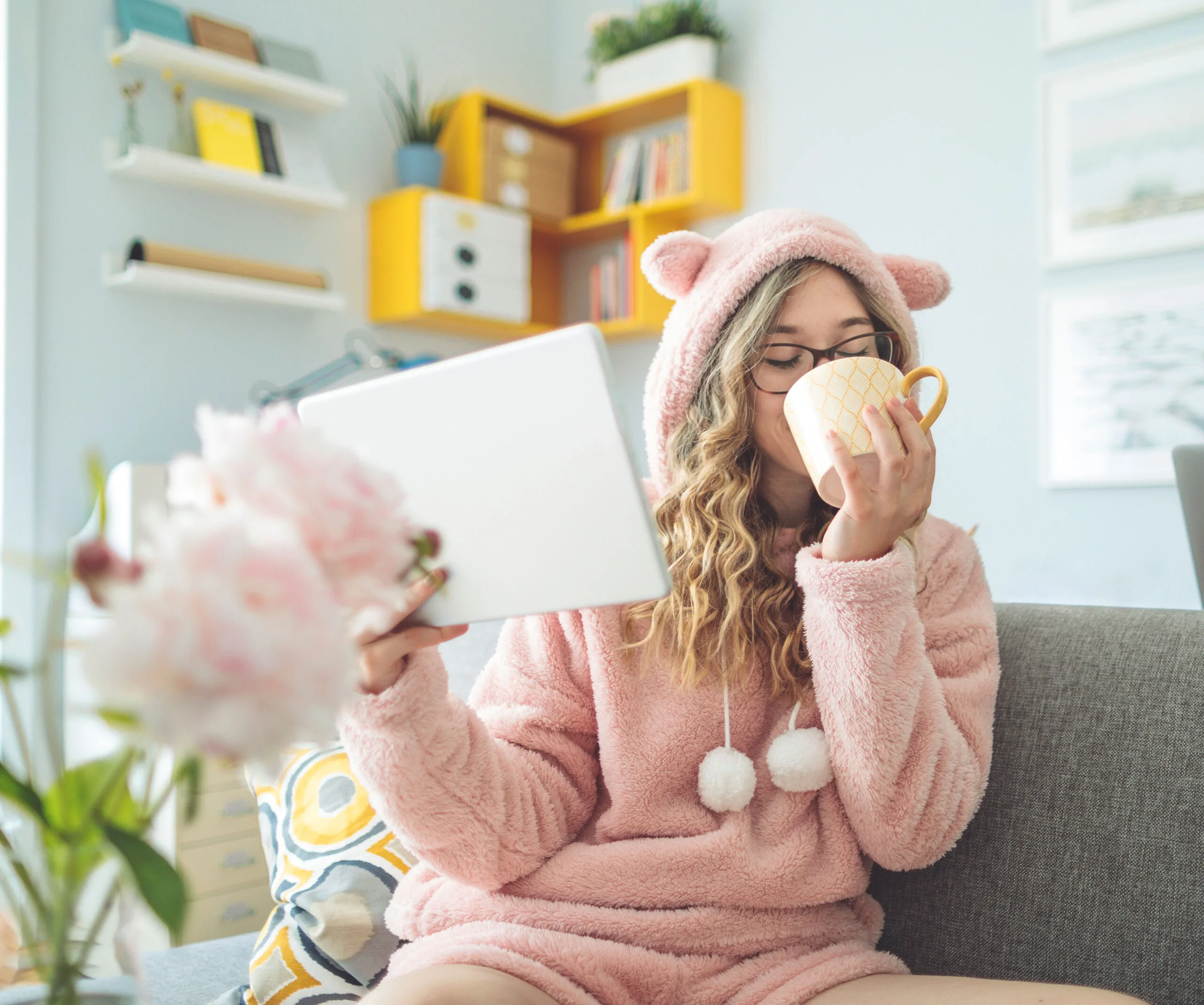 Woman in pink cozy outfit with bear ears, holding a tablet and sipping from a mug on a sofa.