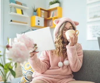 Woman in pink cozy outfit with bear ears, holding a tablet and sipping from a mug on a sofa.