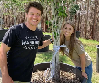Couple smiling next to a tree wrapped with a white ribbon, surrounded by greenery.