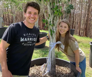 Couple smiling next to a tree wrapped with a white ribbon, surrounded by greenery.