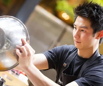 A chef wearing an apron intensely pours contents from a large mixing bowl.