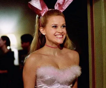 A woman in a playful costume with pink bunny ears and a fluffy top, smiling brightly in a softly lit room.