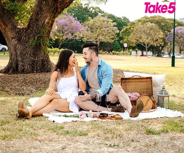 A couple enjoys a picnic in a sunny park, sitting on a blanket with a basket and wine glasses.