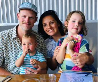 A smiling family of four at a restaurant table, including two young children, one holding a toy.