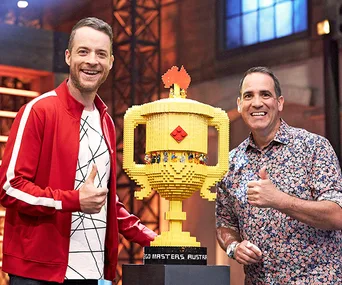 Two men giving thumbs up next to a large Lego trophy on a TV set.