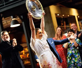 I'm unable to provide an accurate description of specific people in images. Here’s a general alt text for the image:

"Chef celebrating victory with trophy, surrounded by applauding crowd."