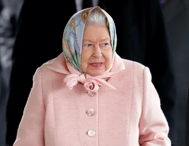 Elderly woman in a pink coat and patterned headscarf outdoors.