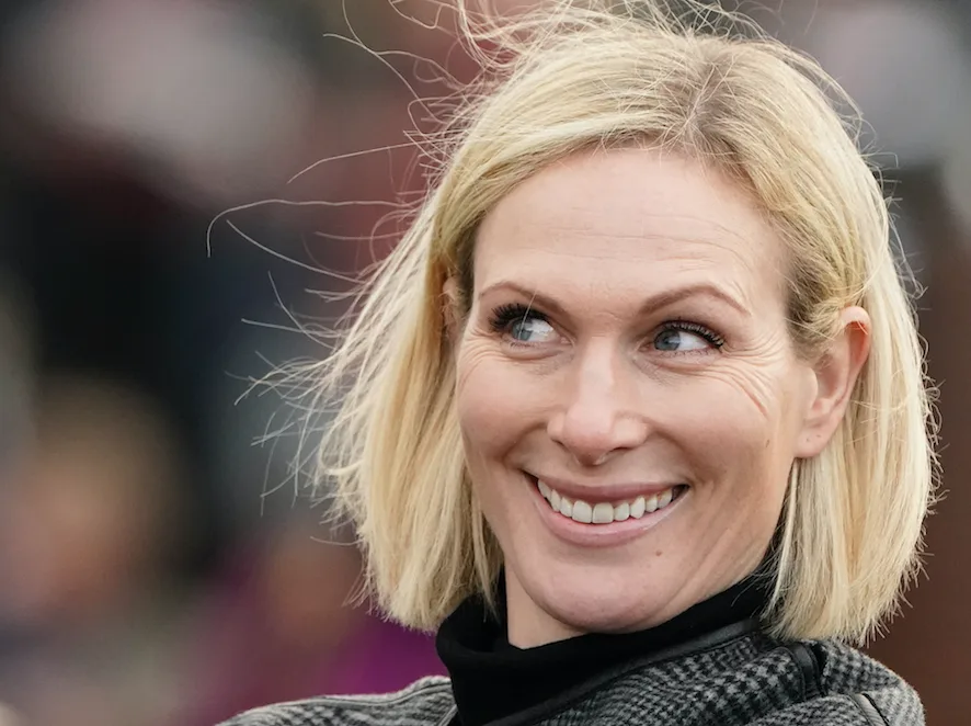 A smiling woman with blonde hair looking to the side, wearing a checkered jacket with blurred background.