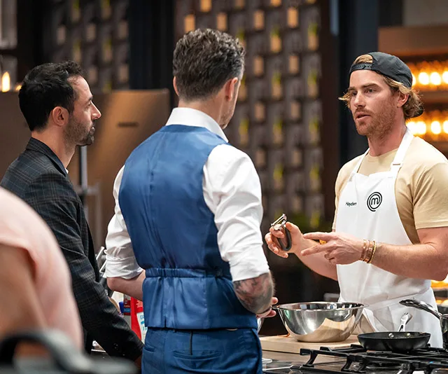 ```html
A MasterChef contestant discusses a dish with two judges in a kitchen setting.
```
