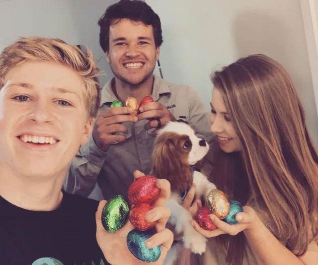 Three people smiling, holding colorful Easter eggs, with a small dog in someone's arms.