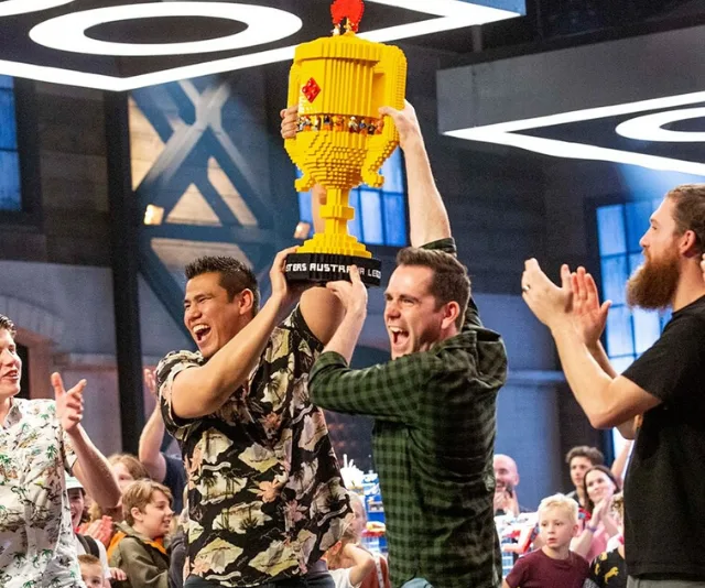 "Two contestants celebrate with a large yellow LEGO trophy on 'LEGO Masters Australia' set, surrounded by cheering crowd."