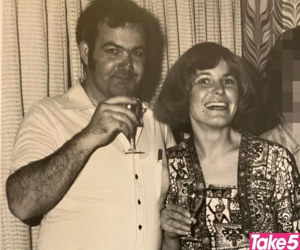 A smiling couple holding drinks in a vintage photo with curtains in the background.