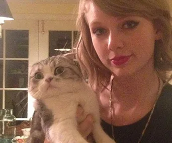 A person holding a surprised-looking cat indoors with a window in the background.