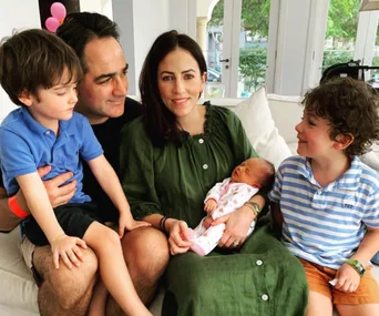 A family of five sits on a couch, with parents holding a newborn baby, surrounded by two young children smiling.