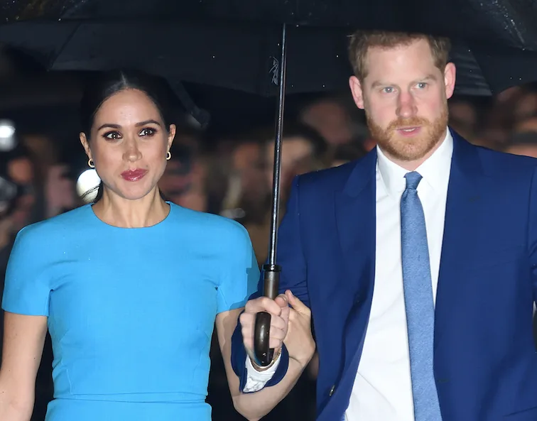 Man and woman under umbrella, dressed in blue outfits, walking in front of a crowd.
