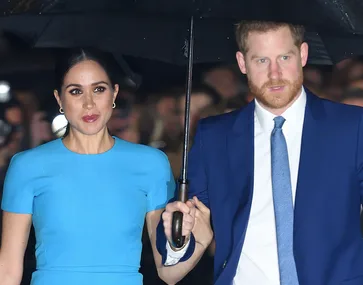 Man and woman under umbrella, dressed in blue outfits, walking in front of a crowd.