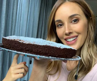 A woman holding a round, flourless chocolate cake topped with powdered sugar, smiling at the camera.