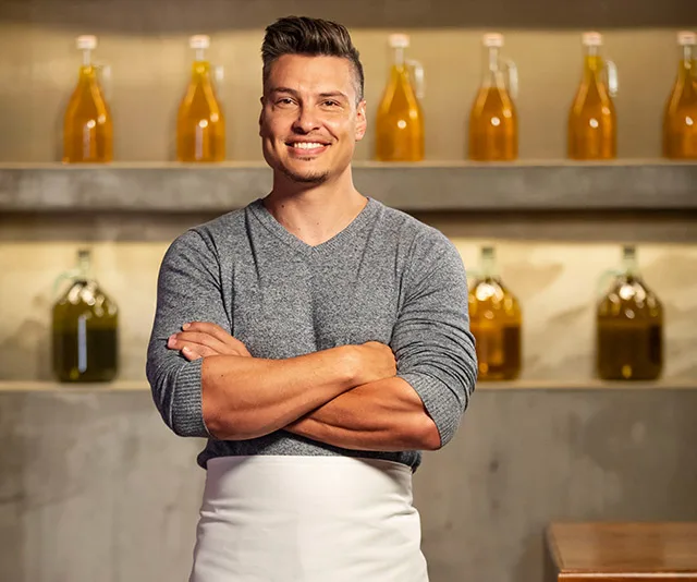 A person in a kitchen setting, wearing a grey sweater and white apron, standing in front of shelves with bottles.
