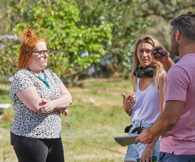 Three people discussing outdoors; one with red hair and glasses, another holding a tool and wearing headphones.