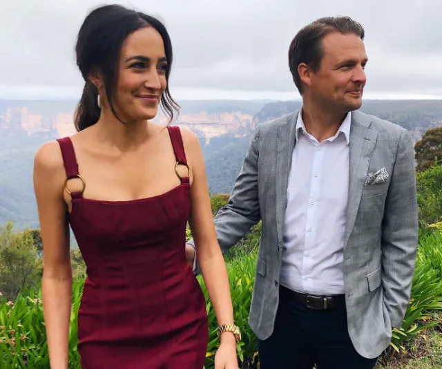 Couple smiling outdoors; woman in maroon dress and man in gray suit, with scenic mountain backdrop.