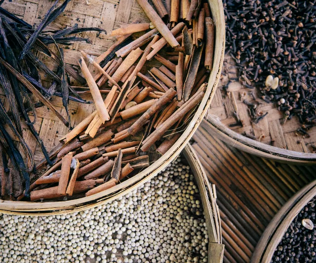 Baskets containing vanilla pods, cinnamon sticks, white pepper, and cloves arranged on a wooden surface.