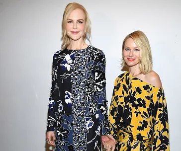 Two women in floral dresses, one in blue and black, the other in yellow and black, holding hands and smiling.