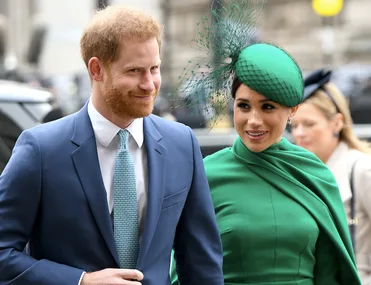 Prince Harry and Meghan Markle smile together, outdoors, Harry in a blue suit, Meghan in green dress and hat.