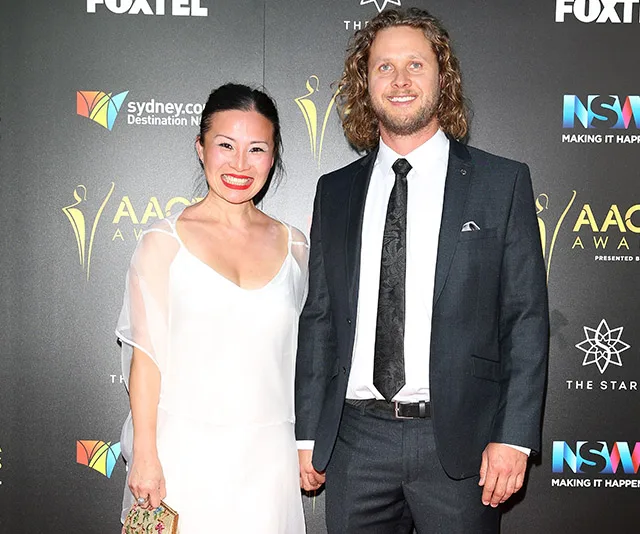 Couple poses on red carpet at AACTA Awards; woman in white dress, man in dark suit.